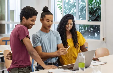 A group of three students standing around a laptop, collaborating and discussing while smiling in a bright indoor environment.