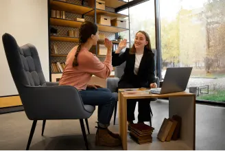 Two women are high-fiving across a table in a modern office setting with bookshelves and a laptop in the background.
