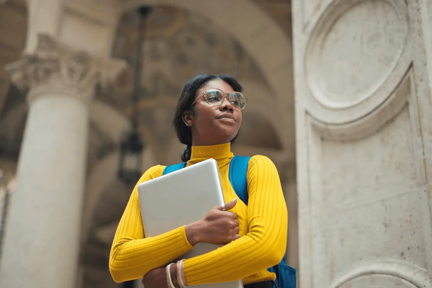 A student wearing a yellow turtleneck sweater and glasses, holding a laptop, with a backpack, standing in front of a historical building.
