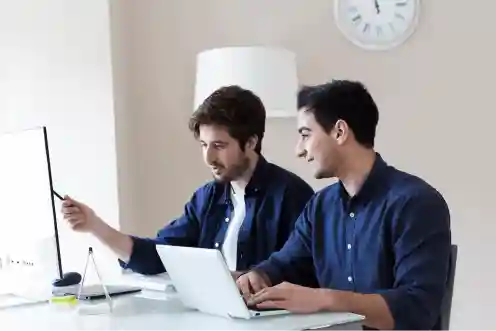 Two people sitting at a desk, one using a laptop and the other pointing at a screen, both dressed in blue shirts.