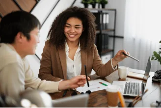 A woman smiling and holding a pen, discussing with a man while sitting at a table with a laptop and papers in an office setting.