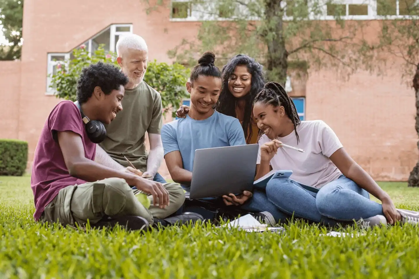 A group of diverse students gathered on the grass, smiling and collaborating around a laptop outdoors.