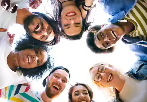 A group of six diverse, smiling individuals, looking up into the camera while standing in a circle