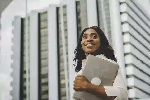 Smiling woman standing outside a building, holding a laptop, dressed in a white outfit with her hair down."
